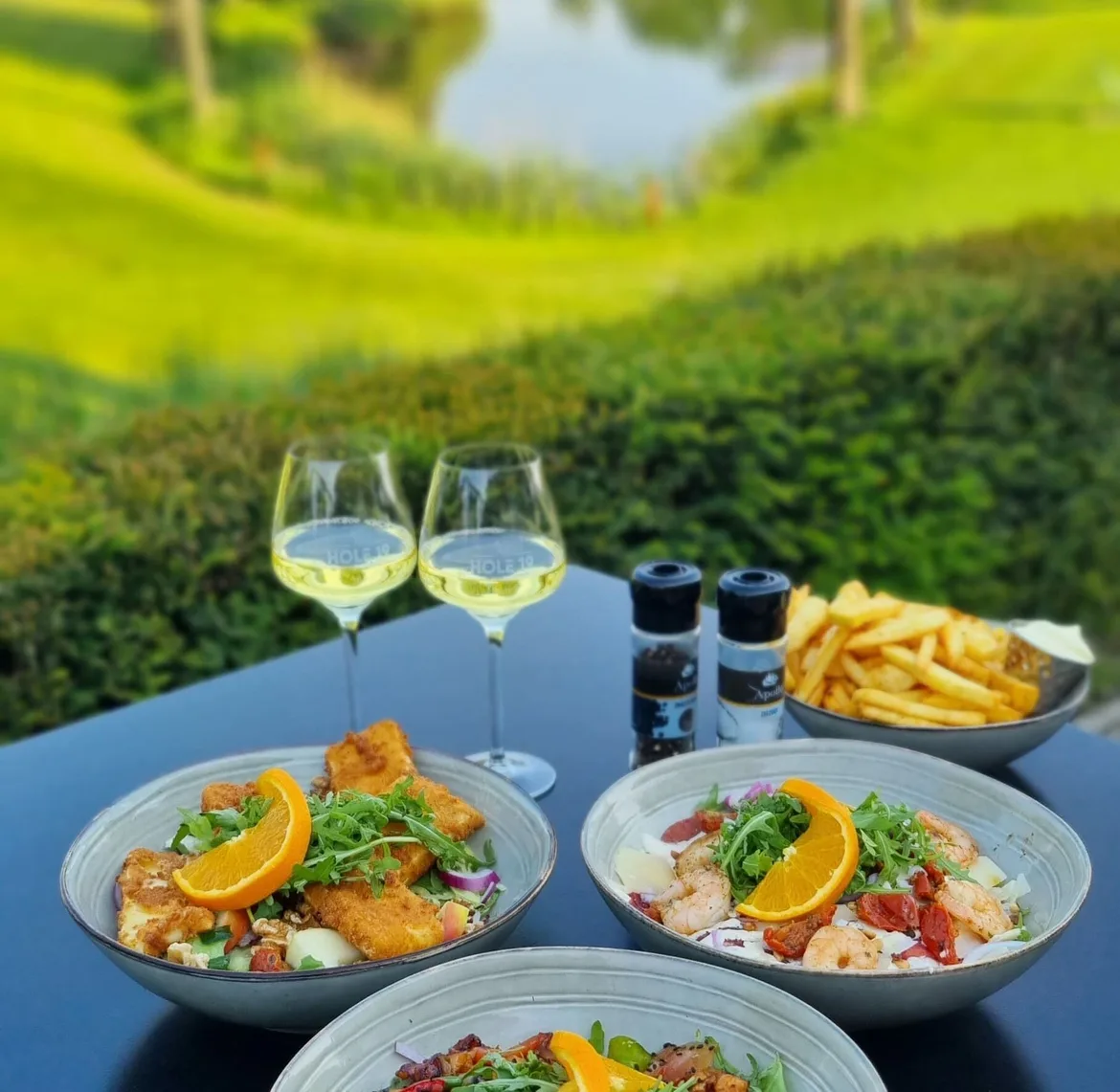 tafeltje met uitzicht op de golfbaan, borden maaltijdsalade, 2 glazen witte wijn en een schaaltje friet op tafel