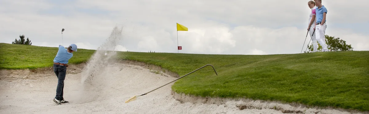 Jongen slaat bal uit bunker de green van hole 16 op. Twee jongeren kijken toe.